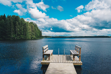 Two wooden chairs bench on a wood pier overlooking a blue lake water with green forest and cloud sky
