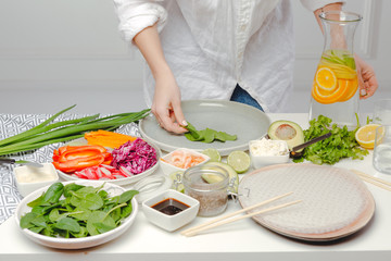 The process of cooking spring rolls - woman's hands putting basil on rice paper. Table with spring rolls ingredients and water with oranges.