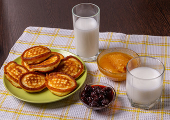Pancakes on a plate, milk in a glass, jam and honey on a napkin on the table.
