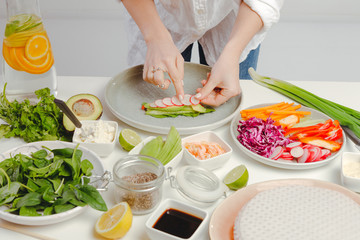 Woman's hands preparing spring rolls, adding sliced radish, basil, cucumbers and seeds in rice paper on kitchen table.