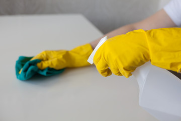 Clean up the kitchen. Woman in yellow rubber gloves spraying cleaner on white table. Selective focus