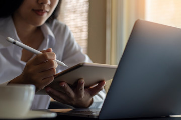 Young asian woman hand holding electronic or stylus pencil and using digital tablet, work on laptop computer with cup of coffee on the desk at home. Online learning, home school or remote work concept