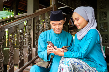 Young couple of malay muslim in traditional costume watching online content in a smart phone during Eid al-Fitr celebration at wooden stair.