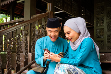 Young couple of malay muslim in traditional costume watching online content in a smart phone with...