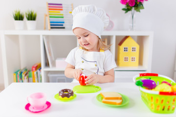 Little blonde girl in white cook uniform playing with toy fruits and vegetables at home, in kindergaten or preschool. Game activities to play with a child at home