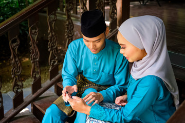 Young couple of malay muslim in traditional costume watching online content in a smart phone during Eid al-Fitr celebration at wooden stair.