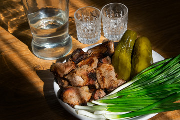 Grilled meat, barbecue, on a white plate with young green onions and salted cucumbers. Bottle of moonshine and crystal glasses, shadow. On wooden table background