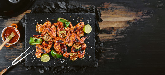 barbecue fried chicken wings on a stone tray lying on a black wooden table with a meat fork. next to a bowl with sauce and brush to cover the wings with sauce