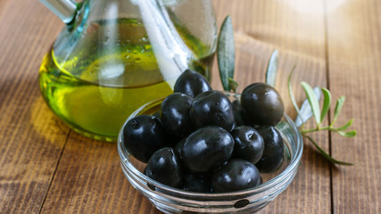 Black canned olives in glass transparent bowl, olive oil bottle in sunlight on the wooden table. Healthy mediterranean food.