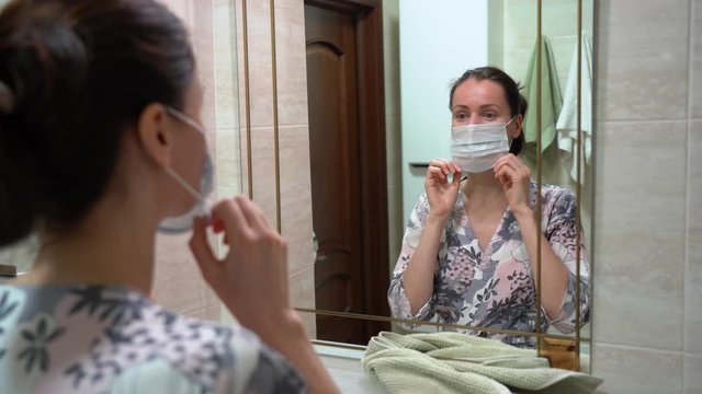 Beautiful Caucasian brunette woman in pajamas put on a protective medical face mask, smile, laugh, look and admire herself in front of the mirror in bathroom at home. Covid19 virus isolation period.