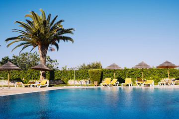 Beautiful tropical beach front hotel resort with swimming pool, sun-loungers and palm trees during a warm sunny day.