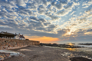 Sunrise at La Rocque harbour, Jersey CI, with stoney beach, sea wall and cloudy sky