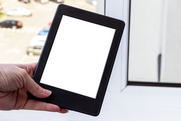 A modern black electronic book with a white blank screen in female hand against a blurred window background. Mockup tablet closeup