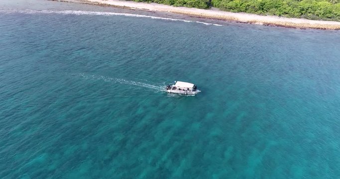 Amazing drone shot of a small speed boat in the Tuheiava Pass in Tikehau, French Polynesia. A small island with palm trees and pink sand flank the lagoon. Coral reef breaks in the distance