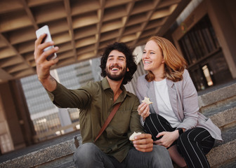 Happy professional couple taking selfie using smartphone while eating sandwich for lunch during break