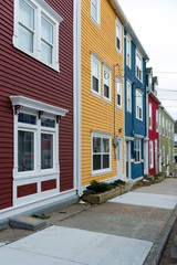 A sidewalk view of a row of colourful wooden houses. The buildings are wooden with windows and doors. There's a concrete sidewalk in front of the buildings.