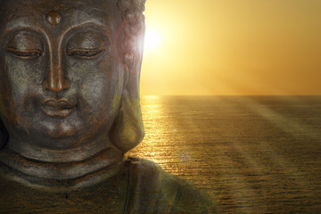 Gesicht einer Buddha-Figur im Gegenlicht eines Sonnenuntergangs am Meer mit Lichtstrahlen und...