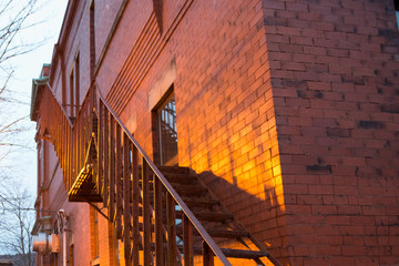 Exterior corner walls of a brown brick building with metal fire escape stairs.  The sun reflecting on the bricks casting a yellow glow. There are glass windows and a metal door in the building. 