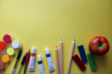 Top view of school stationery: paints, colored pencils, pens, plasticine and apple on a yellow background. Back to school flat lay. Copy space for your text