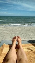 Point of view of young woman lying on deck chair by the sea and tanning. Female legs on chaise-longue relaxing and enjoying during summer vacation on sea. Foot of girl on a resort. Close up Pov.