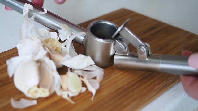 Female hands put fresh garlic into the metalic masher for crushing on wooden tabletop for increased immunity. Healthy food for fighting viruses. Concept of coronavirus or COVID-19 pandemic Close up
