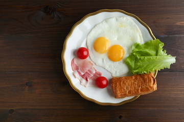 two fried eggs, bacon, green salad and bread are on the plate on dark wooden background flat lay with copy space