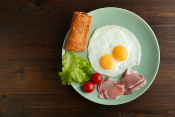 two fried eggs, bacon, green salad and bread are on the plate on dark wooden background flat lay with copy space