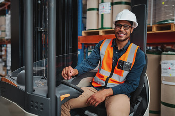 Smiling forklift driver with spectacles sitting in vehicle in warehouse looking at camera wearing a white helmet and safety vest © StratfordProductions