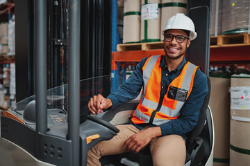 Smiling forklift driver with spectacles sitting in vehicle in warehouse looking at camera wearing a...