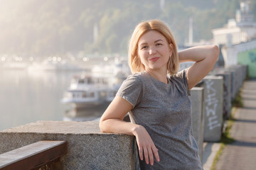 Young blonde woman enjoying city view from the bridge. Lovely woman spending time on the river embankment at sunset. Soft backlit. Lifestyle relax recreation weekend concept