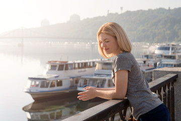 Fototapeta na wymiar Young blonde woman enjoying city view from the bridge. Lovely woman spending time on the river embankment at sunset. Soft backlit. Lifestyle relax recreation weekend concept