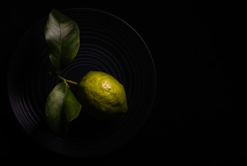 
A green lemon with its black leaves on a black plate. Dark background. Horizontal format, overhead...