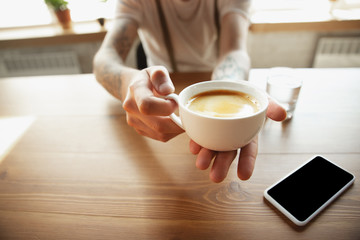 Close up of male hands proposing cup of coffee, sitting at the table with smartphone. Surfing, online shopping, working. Education, freelance, art and business concept. Drinking. Hot aroma drink.