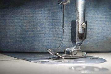 Needle presser foot with thread of white industrial sewing machine with blue jeans close-up.