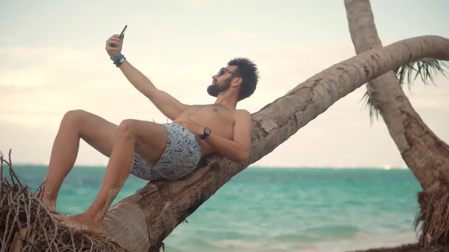 Attractive Tanned Man Sunbathing Using Mobile App For Photo Video.Selfie On Smartphone In Beautiful Place.Man Using Mobile Phone For Selfie Photo.Guy Taking Picture On Vacation Holiday Carribean Beach