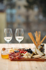 Cheese plate with prosciutto, tomatoes, grapes, olives, pear, grissini, jam, honey and two glasses of wine on a wooden board.