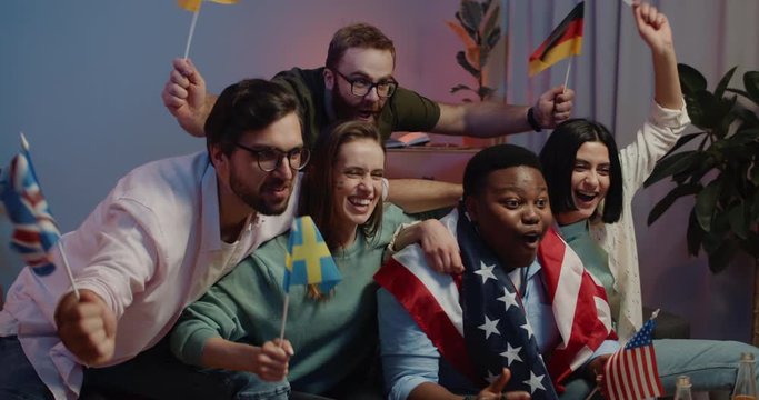 Excited friends with flags having good time together while watching sport championship. Group of smiling young people cheering, shouting and embracing while sitting on sofa