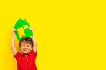 stay home. A boy, a curly laughing emotional kid holds a cardboard house of green color above his head at the top on a bright yellow background, studio. coronavirus pandemic.