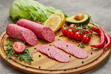 Traditional thinly sliced salami on a wooden cuttingboard  with spices, lettuce, avocado and tomatoes. Cold meat plate