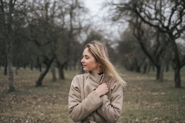 Young beautiful woman in a beige coat walks alone in a park on the street. Resting, windy weather, positive emotions, power balance. Stylish clothes for autumn, spring.