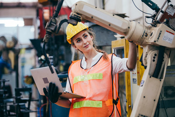 Happy female industrial engineer or technician worker in hard helmet and uniform using laptop checking on robotic arm machine. woman work in heavy technology invention industry manufacturing factory