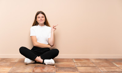 Ukrainian teenager girl sitting on the floor pointing finger to the side