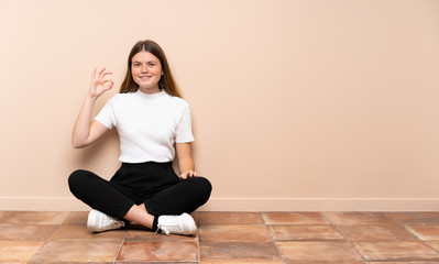Ukrainian teenager girl sitting on the floor showing ok sign with fingers