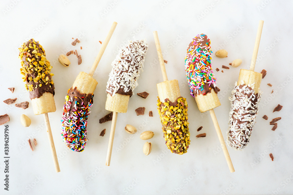 Poster frozen chocolate dipped banana pops with nuts, sprinkles and coconut. top view scattered on a white 
