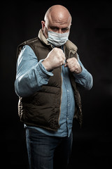 Bald adult man in a medical mask in a fighting stance. Unemployment and self-isolation during the coronavirus pandemic. Black background. Vertical.