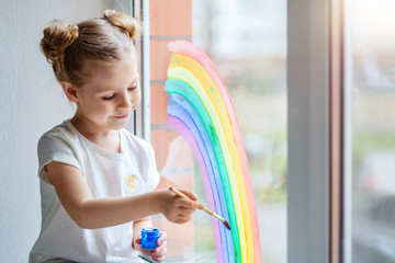 A little girl with blond hair draws a rainbow on the glass with paints. 