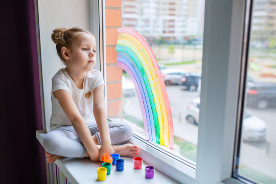 A little girl with blond hair is sitting in front of the window. The child is sad and looks out the window.