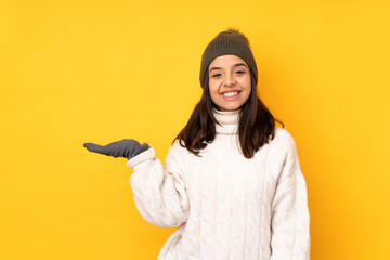 Young woman with winter hat over isolated yellow background holding copyspace imaginary on the palm to insert an ad