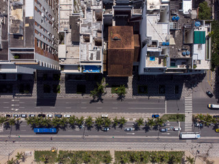Avenue in Ipanema, Rio de Janeiro, seen from top down with palm trees between the roads and along the beach with high rise buildings casting a dark shadow on the sidewalk