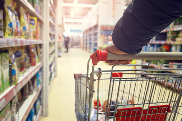 Man with a basket walks in a supermarket. Hand and part of the basket in focus, blurred background